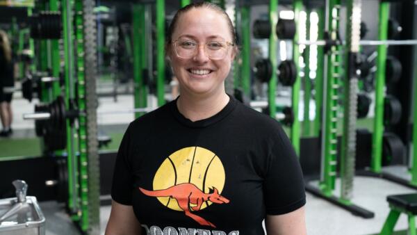 Beth, a member of Carbon Gym, smiling in front of state-of-the-art gym equipment. She wears a "Boomers" t-shirt, representing her story of community, fitness, and wellness at Carbon Gym in Geelong.
