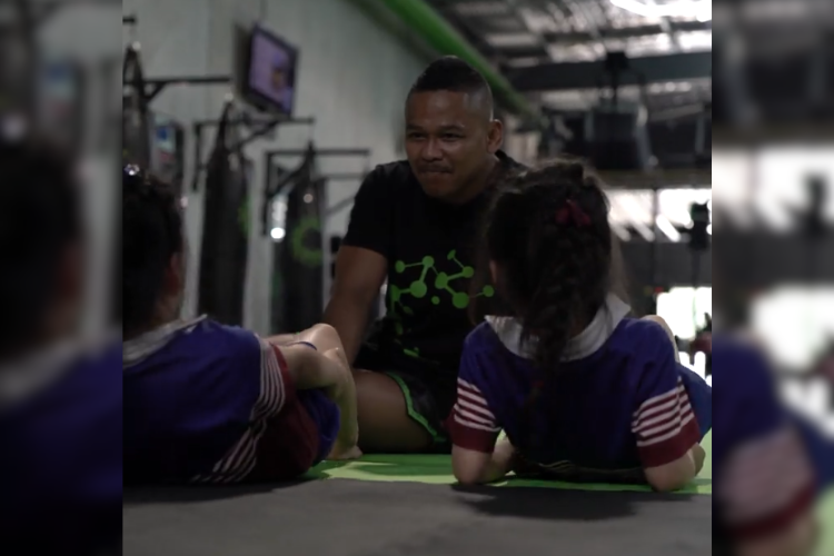 Coach in a black shirt with green details works with two young girls in a kids' martial arts class at Carbon Gym in Geelong. The gym setting includes punching bags and equipment, highlighting a fun and engaging training environment.