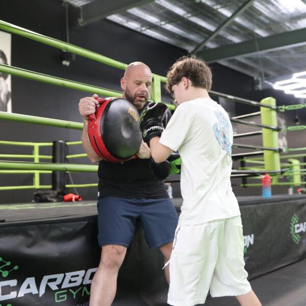 Coach holding focus pads for a teenage boxer in the ring during a teen boxing class at Carbon Gym Geelong