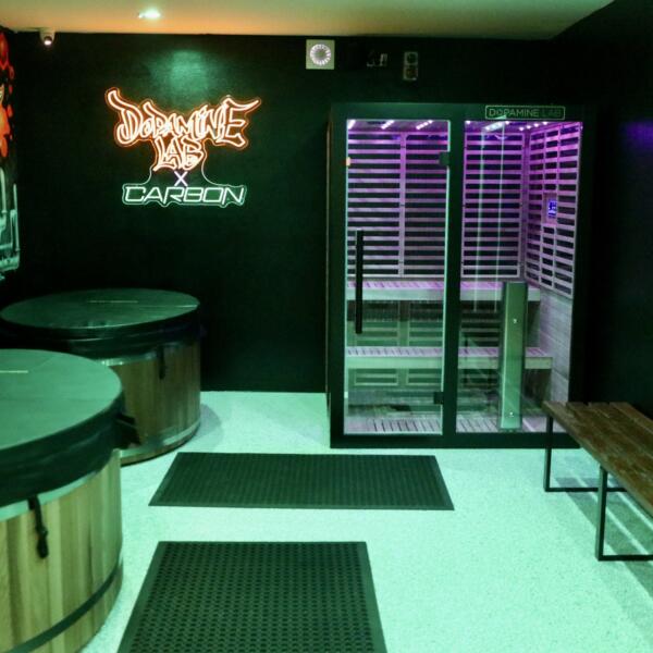 Vertical shot of a recovery room in green ambient light, showing two cold plunge tubs, a purple-lit sauna, and a wooden bench.