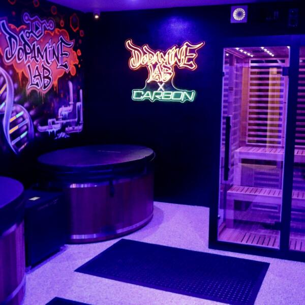 Recovery room lit in deep purple tones, showing a glowing infrared sauna, two wooden cold plunge tubs, and a neon Dopamine Lab sign.
