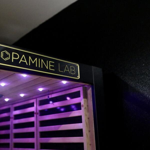 Close-up of the gold Dopamine Lab plaque mounted above a cedar sauna doorway, with purple LED lights glowing inside through the wooden slats.