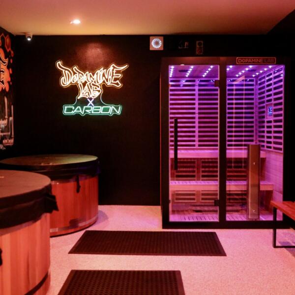 Wide shot of a recovery room featuring a purple-lit infrared sauna, two wooden cold plunge tubs, and a neon Dopamine Lab sign on the wall.