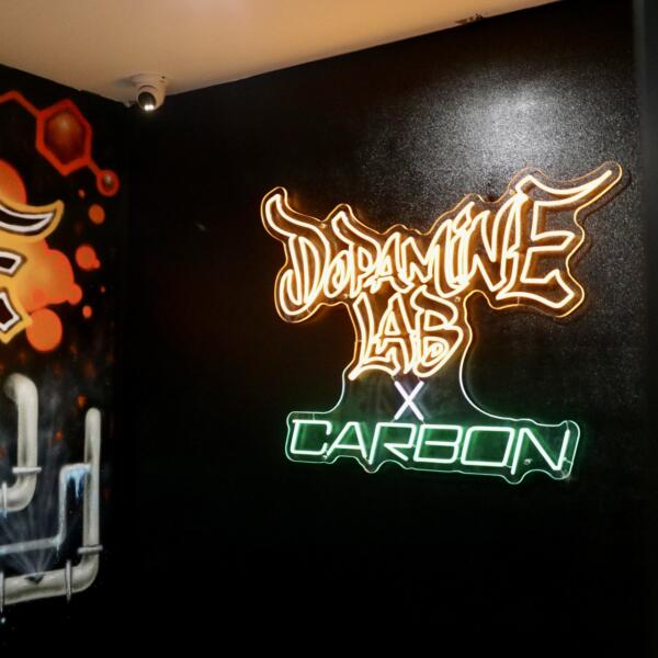 Wide view of an orange and green neon sign reading Dopamine Lab x Carbon on a black wall, next to a painted mural of DNA strands and pipework.