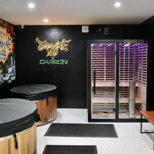 Interior view of a gym recovery room with a purple-lit sauna, two wooden cold plunge tubs, a bench seat, and a Dopamine Lab neon sign on the wall.