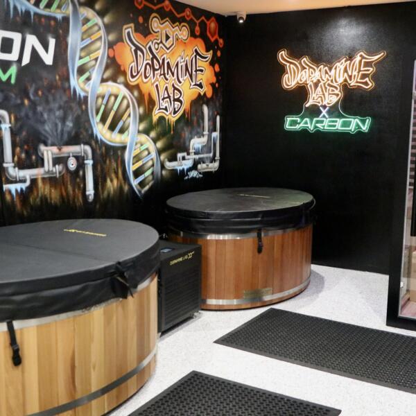 Wide shot of a recovery room with two round wooden cold plunge tubs, a glass-walled infrared sauna glowing purple, and a DNA-themed mural on the wall.