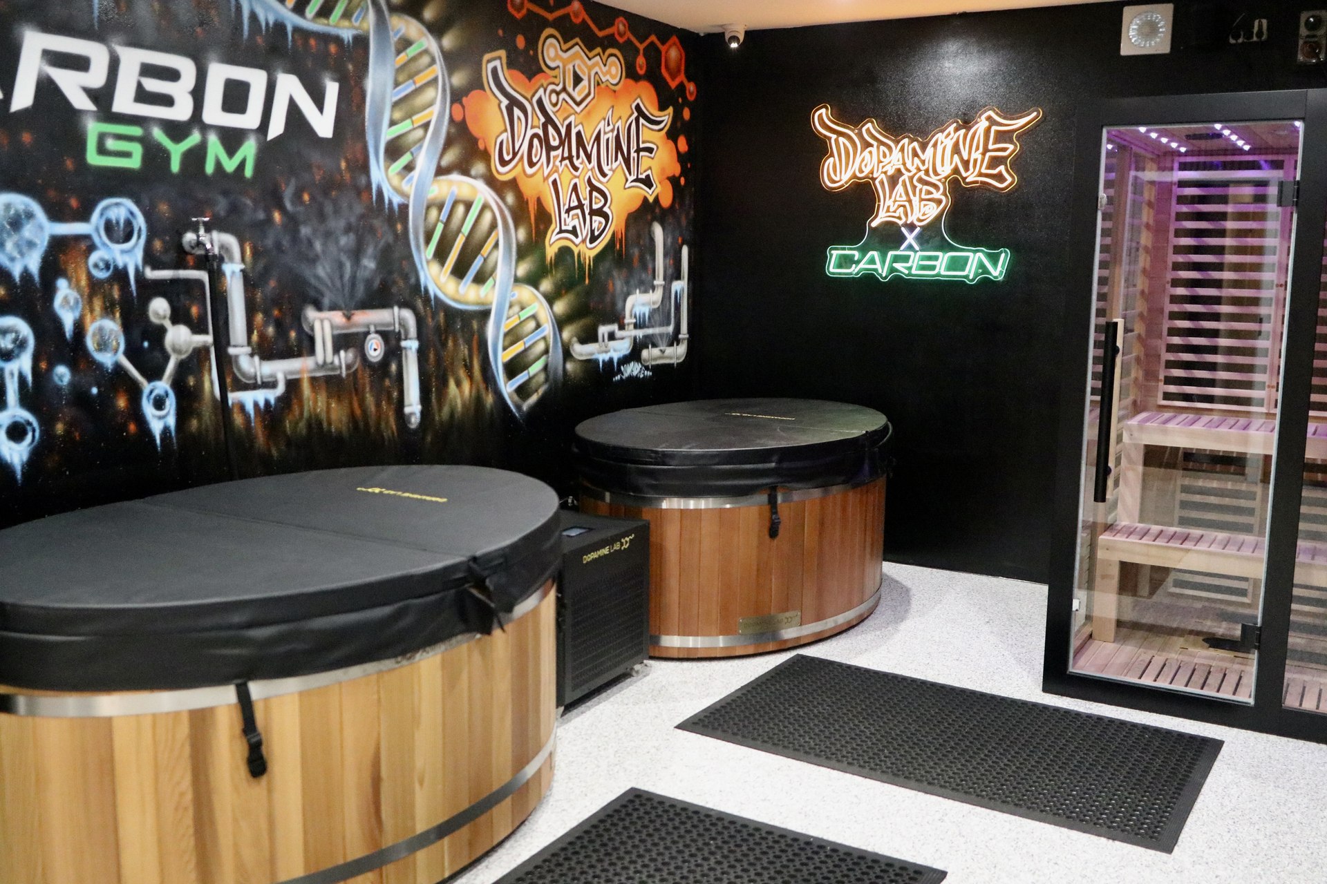 Wide shot of a recovery room with two round wooden cold plunge tubs, a glass-walled infrared sauna glowing purple, and a DNA-themed mural on the wall.