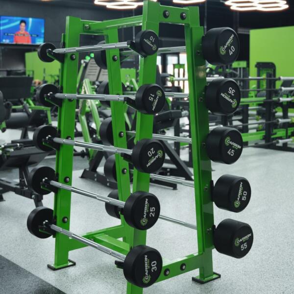 A green metal barbell storage rack loaded with black weight plates, with more racks and equipment visible behind it.