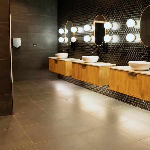 Bathroom interior showing three vessel sinks on a timber vanity, hexagon tiled wall, illuminated round mirrors, and tiled shower entry.