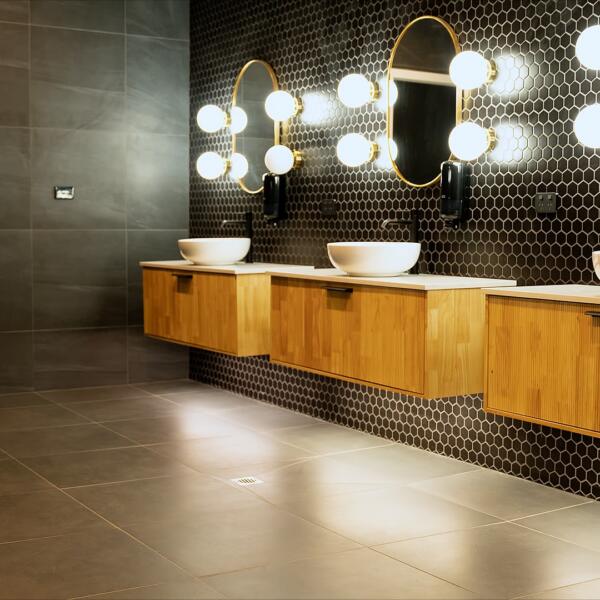 Bathroom interior with three vessel sinks on a wood vanity, hexagon tile wall, round lit mirrors, and a wooden bench near the shower.