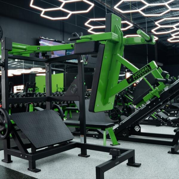 Wide shot of a gym floor with a green and black Arsenal Strength leg press machine, mirrors, dumbbells, and hexagon ceiling lights.