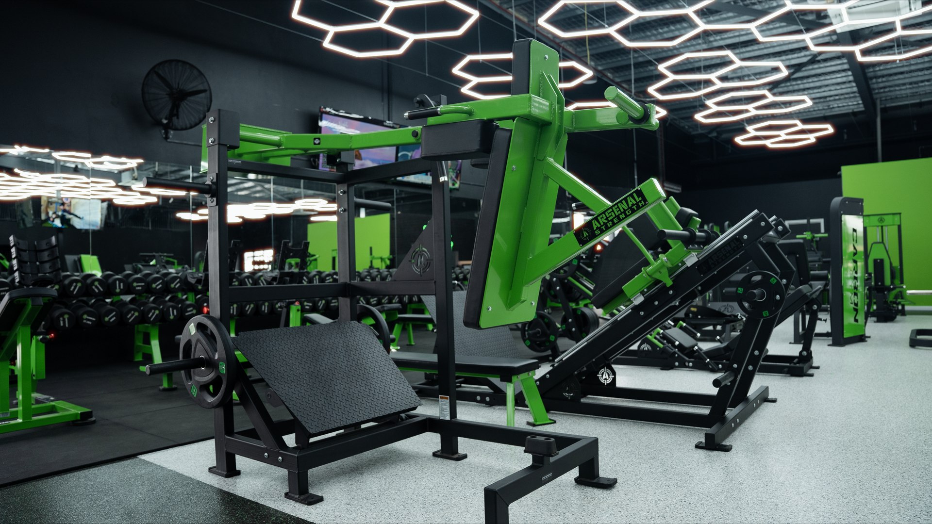 Wide shot of a gym floor with a green and black Arsenal Strength leg press machine, mirrors, dumbbells, and hexagon ceiling lights.