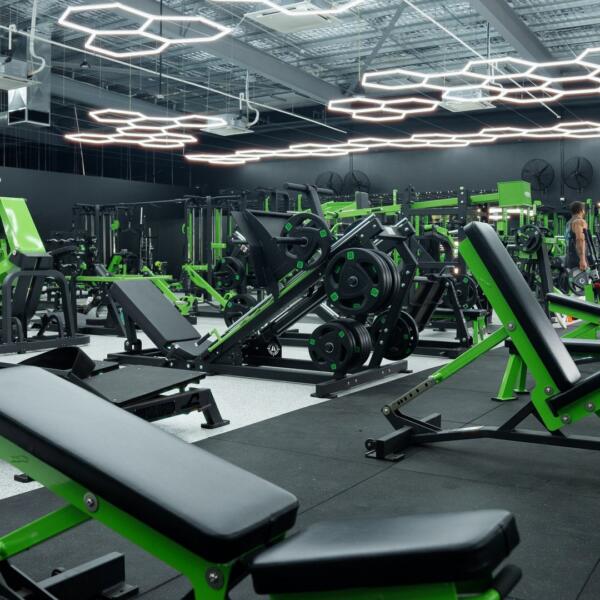 Wide gym floor shot with black and green adjustable benches, cable machines, and hexagon ceiling lights, one person training in the distance.
