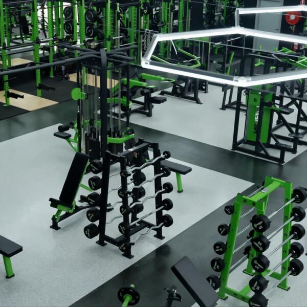 High-angle gym shot of a busy training zone with power racks, benches, and dumbbell storage under bright ceiling lights.