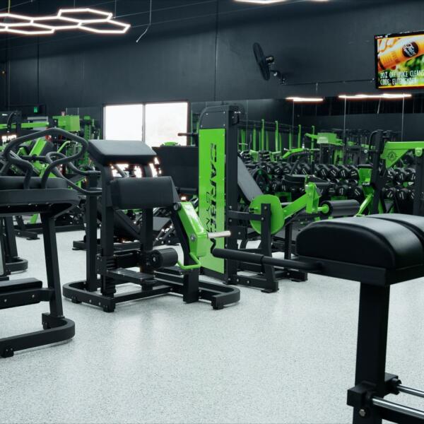 Gym floor with dumbbell racks and strength machines under hexagon ceiling lights, with TV screens mounted on the wall.