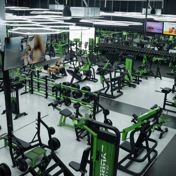 High-angle wide shot of a large gym floor packed with strength machines, benches, and dumbbell racks, with a member training.