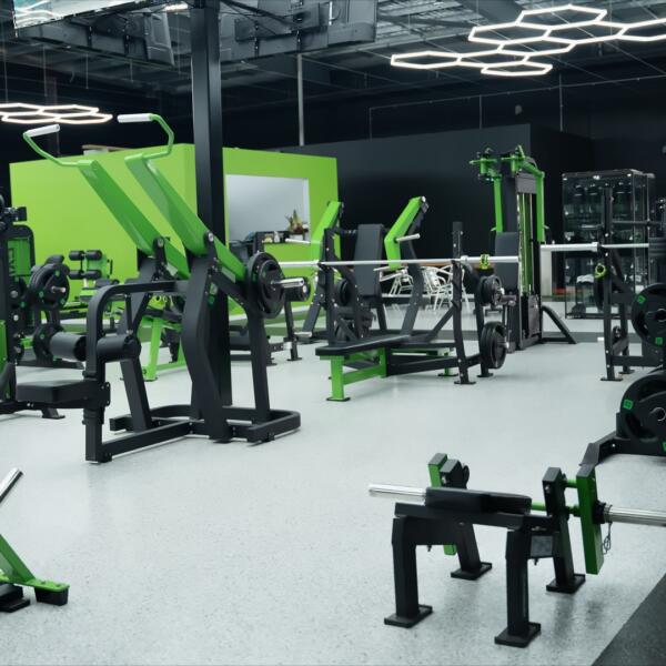 Wide gym interior view with rows of green and black strength machines, benches, and open floor space under bright lighting.