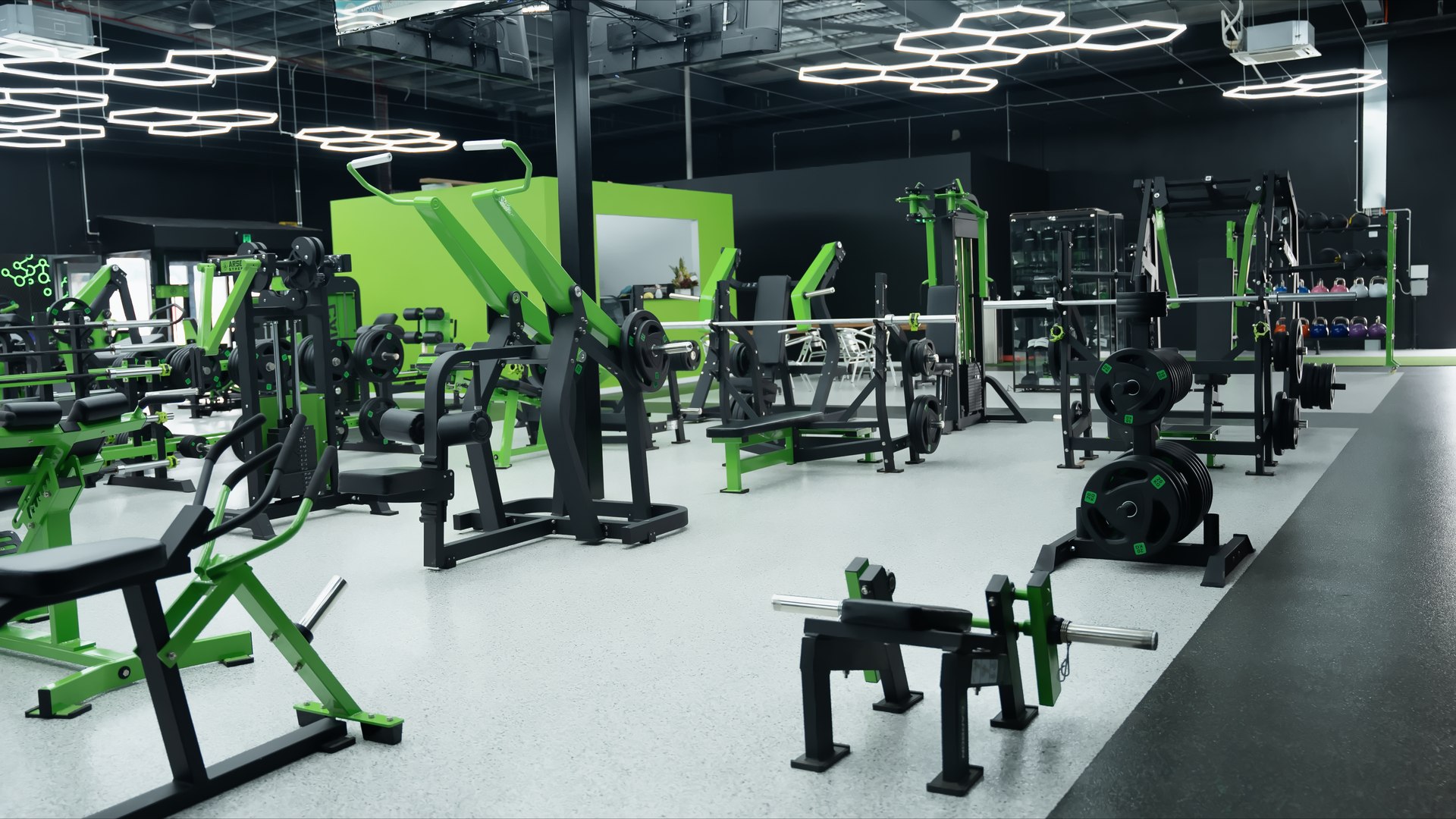 Wide gym interior view with rows of green and black strength machines, benches, and open floor space under bright lighting.