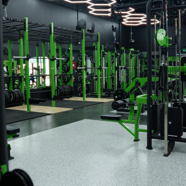 Wide interior shot of a gym floor lined with green power racks and barbells under glowing hexagon ceiling lights.