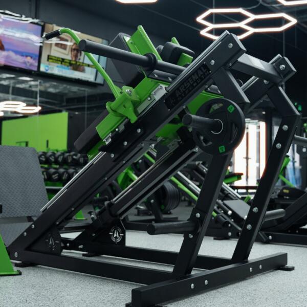 Angled view of a green and black leg press machine on a gym floor, with wall-mounted TVs and hexagon lights above.
