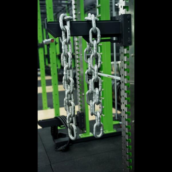 Close-up of two thick metal chains hanging from the crossbar of a green and black power rack in a dark gym.