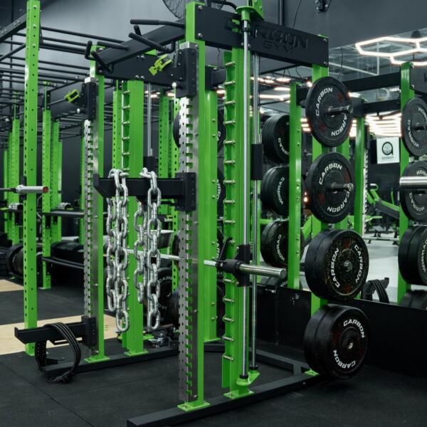 Green and black power rack loaded with weight plates and draped with metal chains, mirrors reflecting the gym behind it.