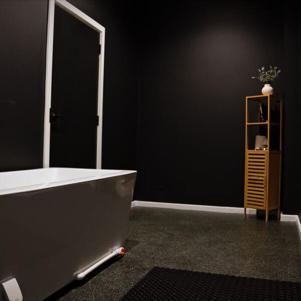 Dark private bathroom interior with a white freestanding bathtub, black walls, white trim, and a timber shelving unit.