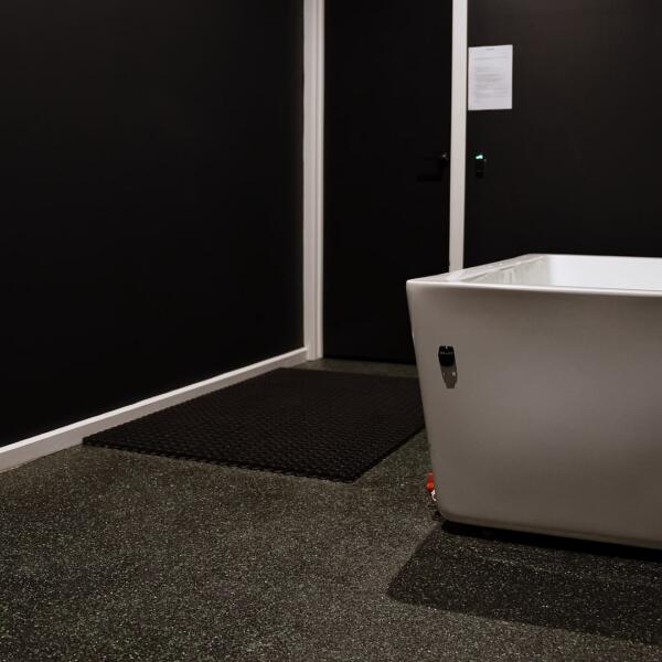 Close view of a white freestanding bathtub in a dark room near an open door, with a timber shelf and black floor mat visible.