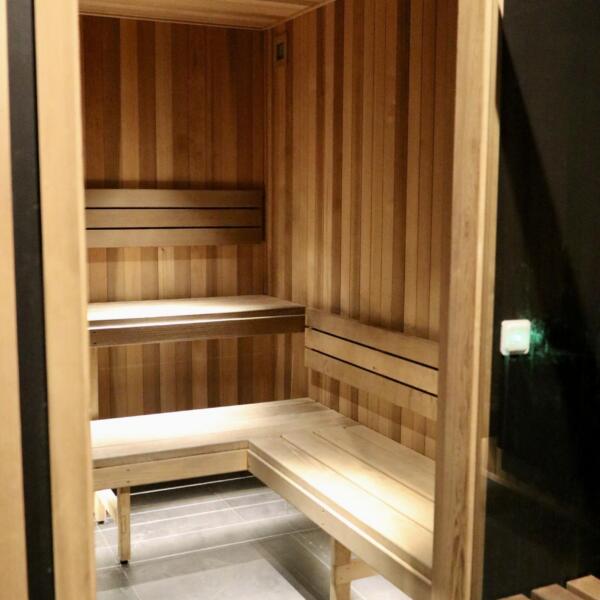 Open glass door of a cedar-lined infrared sauna showing tiered wooden benches and soft warm lighting, dark tiled entry in foreground.