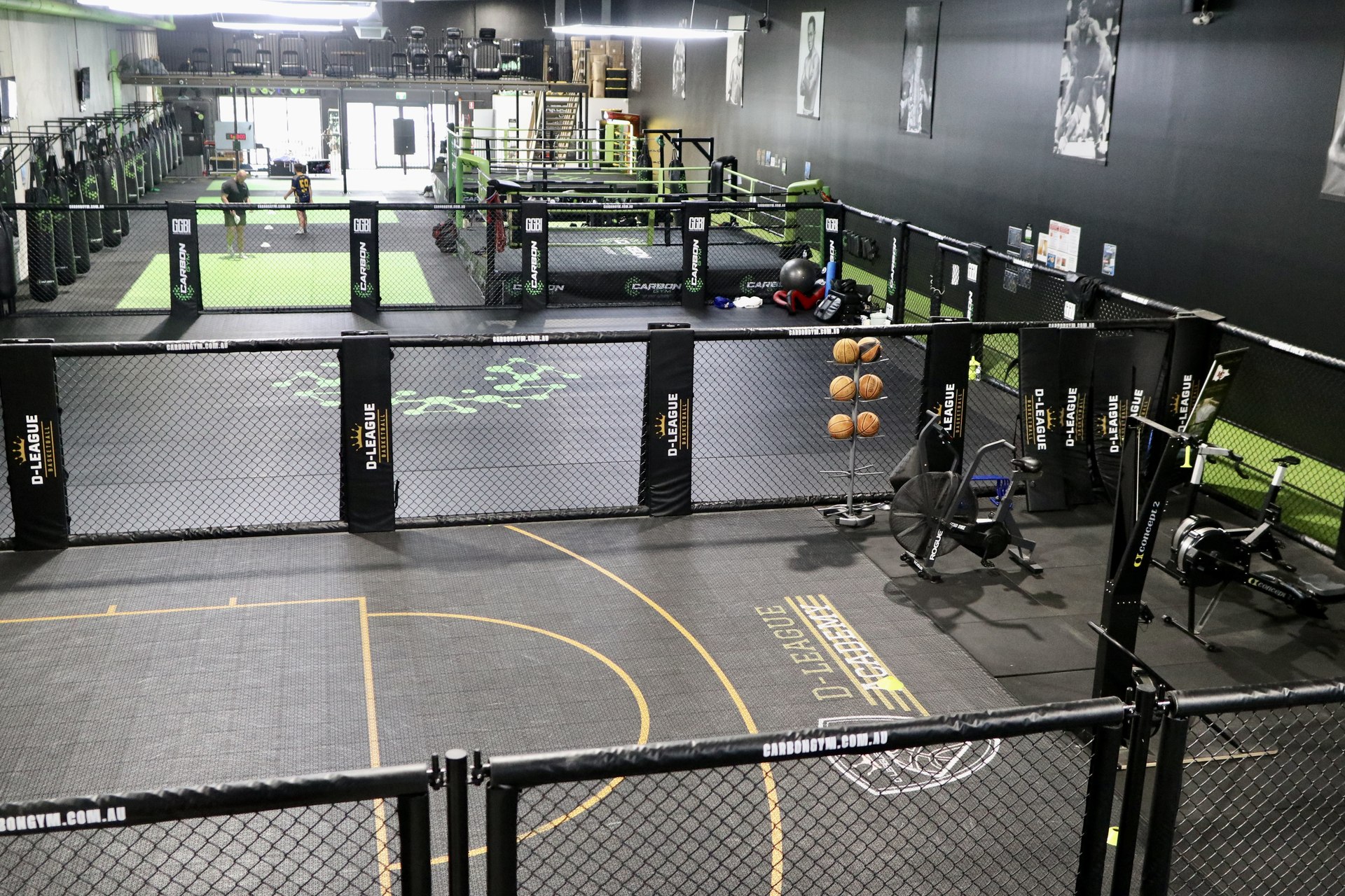 A coach and member training together on a gym mat inside a fenced studio area, with a boxing ring behind them.
