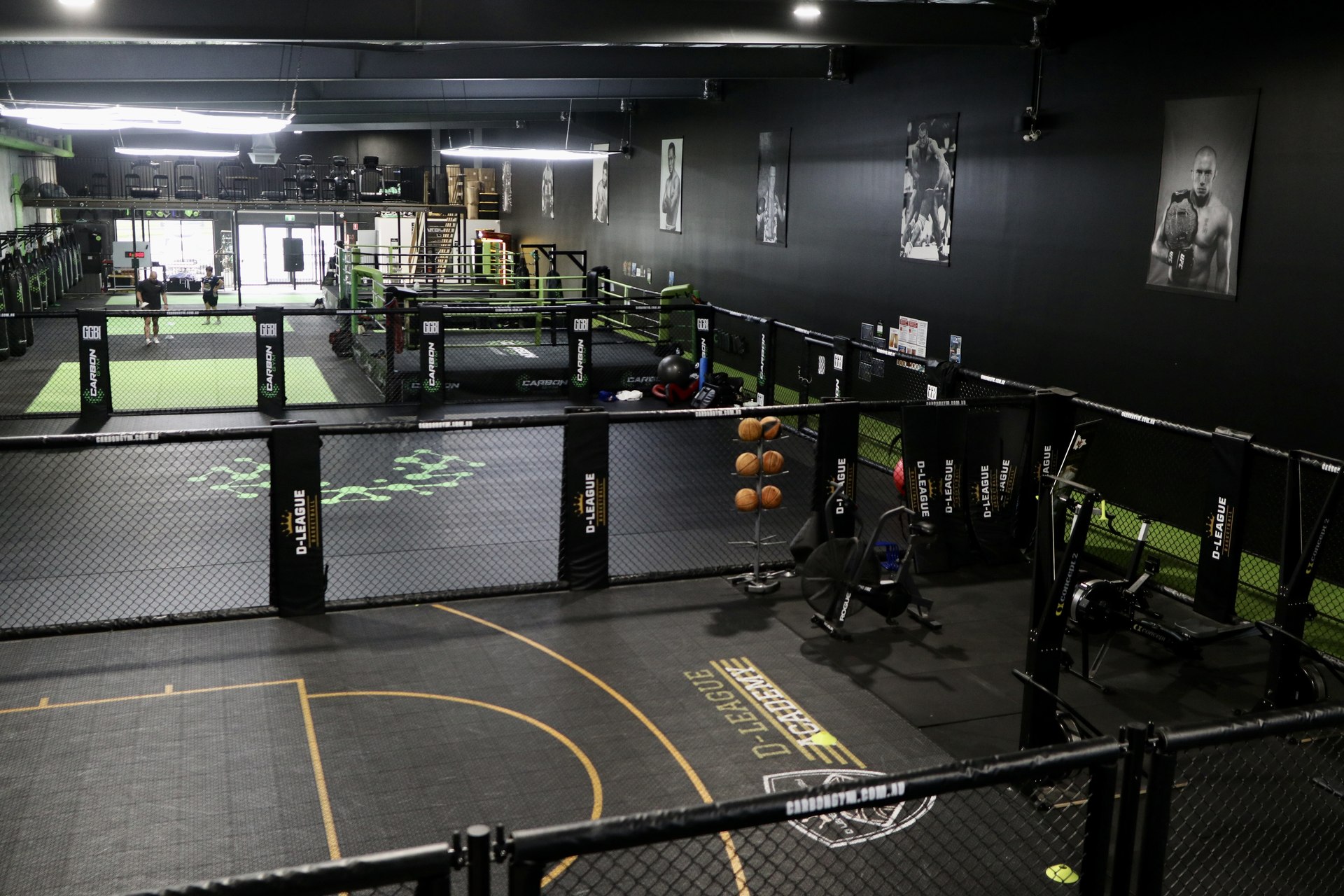 Two gym members training together on green mats inside a caged studio, with a boxing ring visible in the background.
