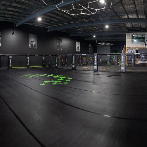 Panoramic photo of an empty gym training floor with a chain-link cage, boxing ring in the distance and large CARBON GYM wall lettering.