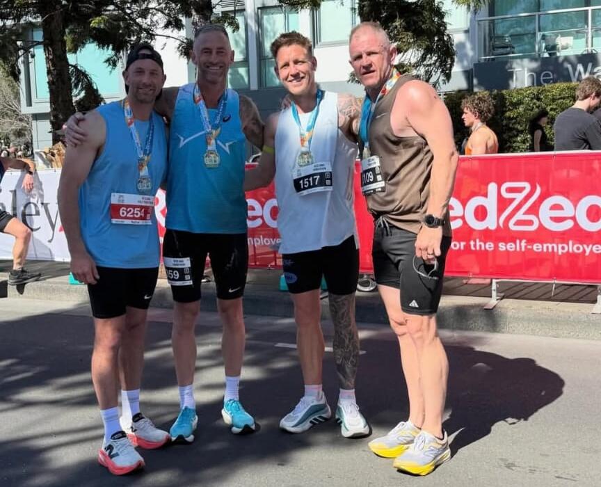 Four runners wearing finisher medals and race bibs stand together at the Geelong Running Festival marathon finish, with Carbon Gym coach Billy Nicholls